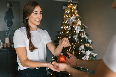 A young and beautiful woman decorates the Christmas tree with ornaments on Christmas Eve. The concept of a festive, cozy home atmosphere. The girl smiles and holds a Christmas ornament in her hands against the backdrop of the tree and lightsの写真素材