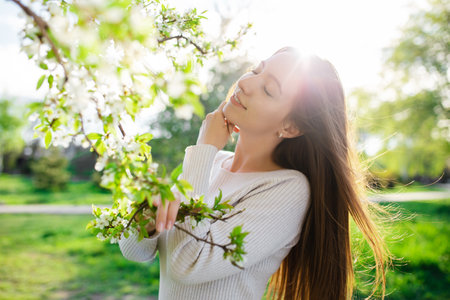 A beautiful young woman is posing, capturing the essence of spring, love, happiness, and femininity in a blossoming spring park at sunset. The setting conveys the joys of the seasonの写真素材