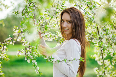 A close-up view captures a lovely girl with flowing long hair, striking a pose amid the blossoming trees of a spring park. The scene embodies the notions of femininity, happiness, and loveの写真素材