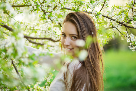 A close-up view captures a lovely girl with flowing long hair, striking a pose amid the blossoming trees of a spring park. The scene embodies the notions of femininity, happiness, and loveの写真素材