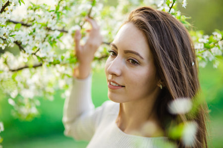 A close-up captures a lovely girl with flowing long hair posing amidst the blossoming trees of a spring park. It embodies the concepts of femininity, happiness, and loveの写真素材