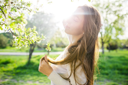 A close-up captures a lovely girl with flowing long hair posing amidst the blossoming trees of a spring park at sunset. It embodies the concepts of femininity, happiness, and loveの写真素材