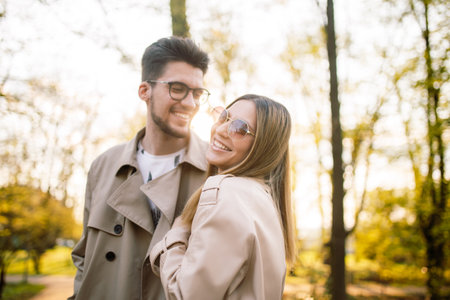 Portrait of a guy and a woman posing and smiling with teeth laughing in beige trench coats in the park at sunset. Spring sunny landscape. Concept of beauty, love and happiness. Springtimeの写真素材