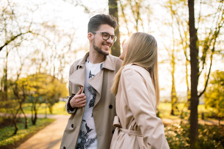 Portrait of a guy and a woman posing and smiling with teeth laughing in beige trench coats in the park at sunset. Spring sunny landscape. Concept of beauty, love and happinessの写真素材