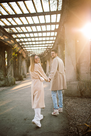A young couple in beige trench coats walks through the park and has fun. A woman and a man pose on an alley on a sunny spring day at sunsetの写真素材