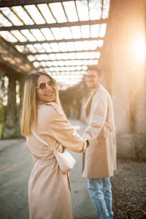 A young couple in beige trench coats walks through the park and has fun. A woman and a man pose on an alley on a sunny spring day at sunsetの写真素材