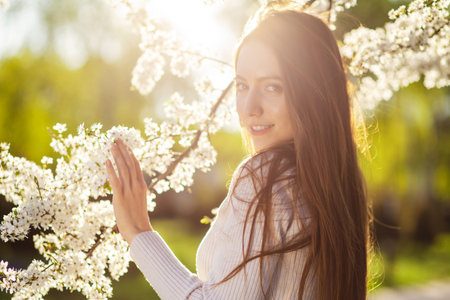 A beautiful long-haired girl in a dress enjoys nature near flowering apple and cherry trees in a spring park at sunset. The concept of happiness, joy, calmness and femininityの写真素材