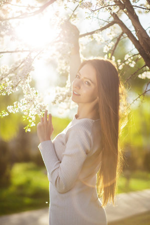 Portrait of a woman in a beautiful dress posing next to a flowering tree in a autumn park at sunset. The concept of femininity, happiness and loveの写真素材