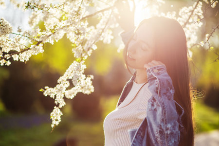 A beautiful young woman with long hair relaxes in a park with flowering trees at sunset in early spring natureの写真素材
