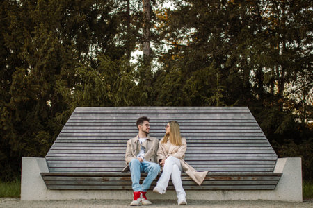 Beautiful stylish young couple sitting on a bench. A woman and a man are talking and enjoying time and spring weather togetherの写真素材