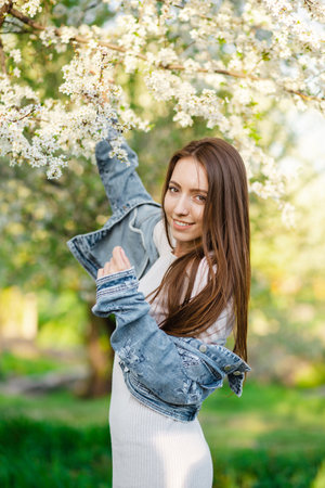 A young woman in a denim jacket and a beautiful dress enjoys a fragrant garden filled with blooming apple and cherry trees in the spring at sunset. The concept of femininity, happiness and loveの写真素材
