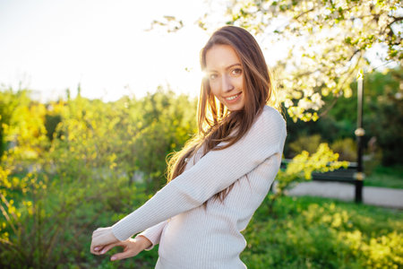 A young woman in a beautiful beige dress is having fun in a blooming garden near a cherry apple tree at sunset. Girl with long hair smiling with teeth in parkの写真素材
