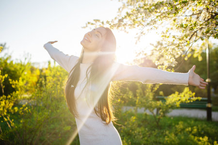 A young woman in a beautiful beige dress is having fun and enjoying herself in a blooming garden near a cherry apple tree at sunsetの写真素材