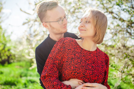 Blond woman and man hug and spend time happily near a blossoming cherry apple tree on a sunny spring day. Concept of love, family and joyの写真素材