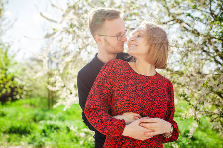 Blond woman and man hug and spend time happily near a blossoming cherry apple tree on a sunny spring day. Concept of love, family and joyの写真素材
