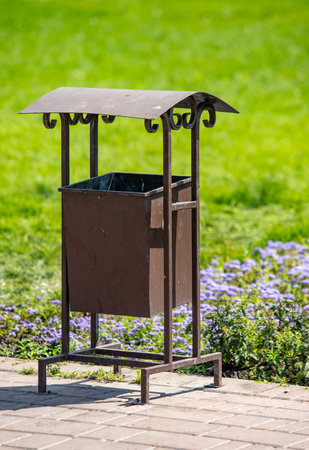 A trash can with a metal roof sits in a grassy area. The trash can is empty and has a lid on top. The area is lush and green, with flowers nearbyの写真素材