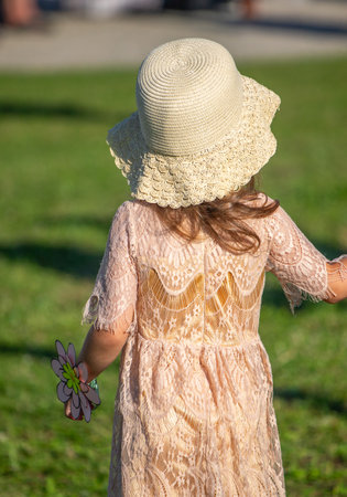 A young girl wearing a white hat and a pink dress is walking in a field. She is holding a flower in her handの写真素材