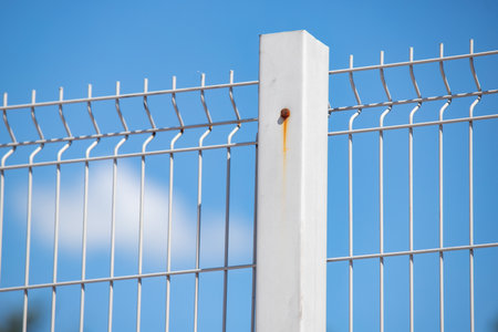 A white metal fence with a rusty post. The fence is surrounded by a blue sky.の写真素材