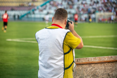 A man wearing a white shirt and yellow vest is taking a picture of a soccer game. The man is standing on the sidelines of the field, and there are several other people in the backgroundの写真素材