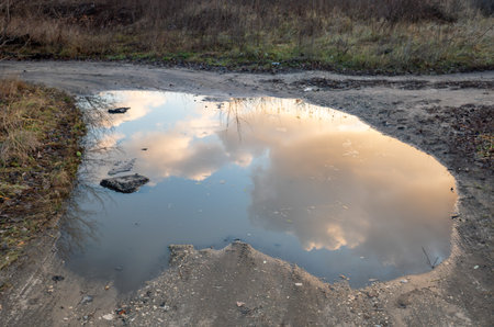 A puddle of water in the middle of a dirt road. The water is reflecting the sky and the cloudsの写真素材