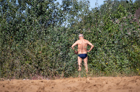 A man is standing in a field of tall grass and bushes. He is wearing a blue swimsuit and he is looking at something in the distanceの写真素材