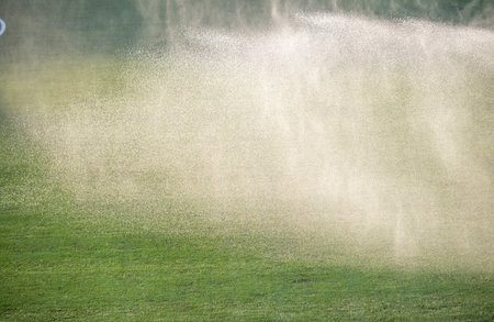 A large white cloud of water is spraying over a green field. The water is coming from a sprinkler system, and it is creating a misty atmosphere. The scene is peaceful and calmingの写真素材