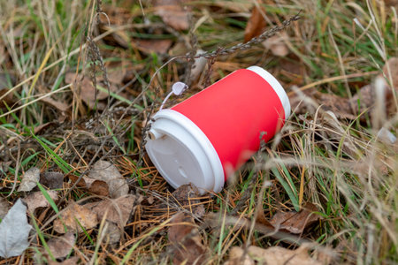 A red cup with a white lid is laying on the ground in a grassy area. The cup is empty and is a disposable coffee cup. The scene suggests a sense of wastefulness and disregard for the environmentの写真素材