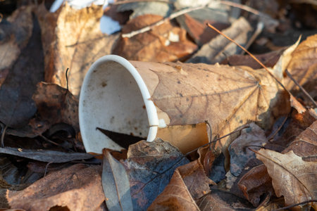 A cup is broken and laying on top of a pile of leaves. The cup is white and has a black handle. The leaves are brown and scattered around the cup. The scene gives off a feeling of abandonmentの写真素材