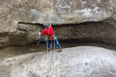 A man in a red shirt is climbing a rock. He is wearing a harness and is using a rope to climb.の写真素材