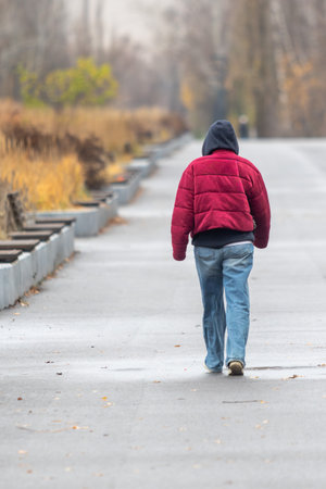 A man in a red jacket and blue jeans walks down a sidewalk. The image has a mood of solitude and lonelinessの写真素材