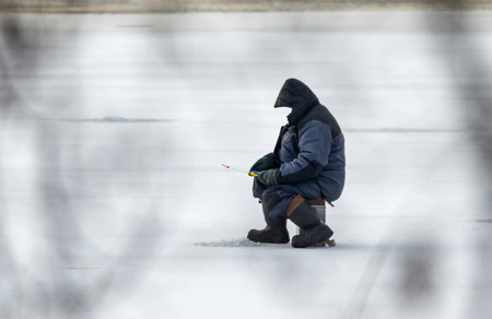 A man sits on a bench in the snow, holding a fishing pole. The scene is quiet and peaceful, with the man alone in the coldの写真素材