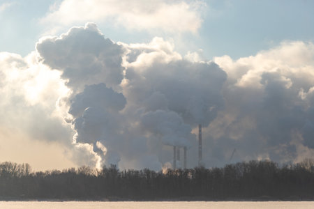 A large cloud of smoke is rising from a factory. The sky is cloudy and the sun is setting