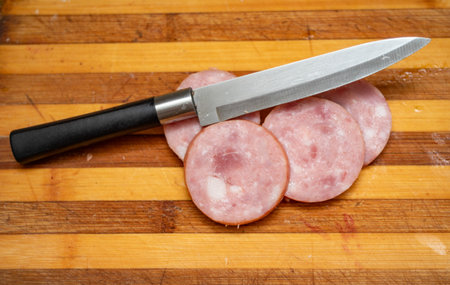 A knife is on a cutting board with a piece of ham. The knife is silver and blackの写真素材