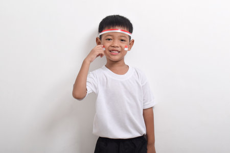 Cute little Asian boy wearing flag headband posing with index finger on cheek isolated on white background. Concept of Indonesia's independence day on August 17の写真素材