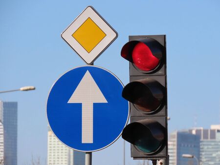 Traffic lights with red signal on and road signs prohibitory Ahead only, Priority road, blue sky and blurred high rise buildings in background, on sunny spring dayの写真素材
