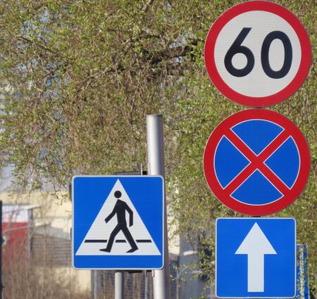 Cluster of road signs: Pedestrian crossing, Straight on only, No waiting, Speed limit 60 km per hour. Background of budding trees on bright spring day. Blue, white, red, black colors の写真素材