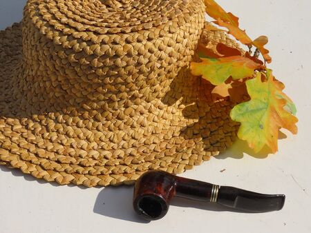 Wooden smoking pipe and rustic straw hat with a branch of oak tree leaves in autumn colours green, brown, red, yellow, gold on off white tableの写真素材