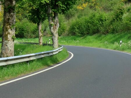 Countryside road without middle divide, with safety barrier towards drop in valley, bright spring day, winding grey tarmac roadの写真素材