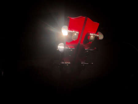 Bright red headstock and shining brass tuning pegs of an acoustic guitar from behind, sun rays on...