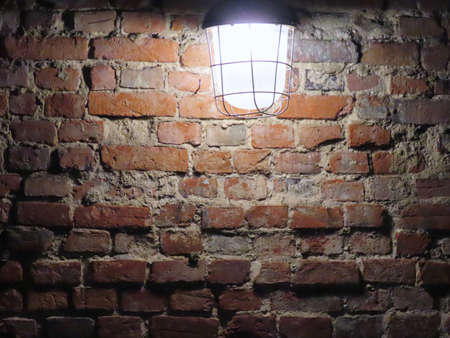 Red brick wall and alight outdoors lamp covered with safety cage, background texture with copy spaceの写真素材
