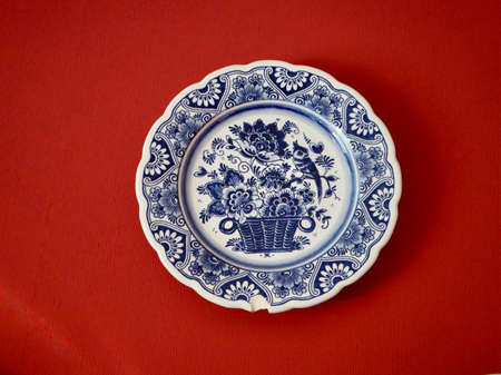 Blue - white delftware with flower bouquet, basket, bird - in centre and ornamental border with defect. Antique Delft plate on red wall. Luxembourg, Luxembourg - December 16, 2021.のeditorial素材