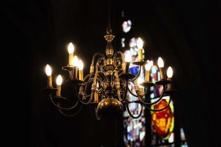 Bronze candelabra with multiple branches, electric light bulbs in shape of candles. Interior detail with blurred out stained glass window in the backgroundの写真素材
