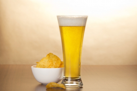 Glass of light amber beer and bowl with chips against brown wallの写真素材
