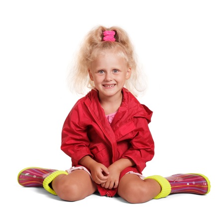 Adorable little blond girl in a red jacket and colorful rubber boots sitting isolated on white background.の写真素材