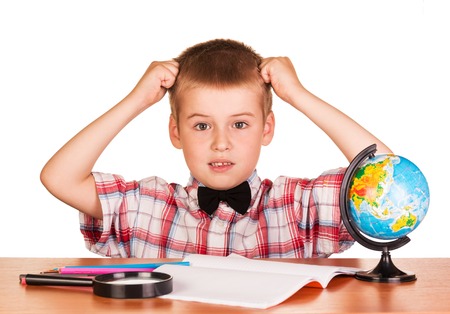 Befuddled boy sitting at a table on a notebook, pencils, magnifying glass and globe isolated on white background.の写真素材