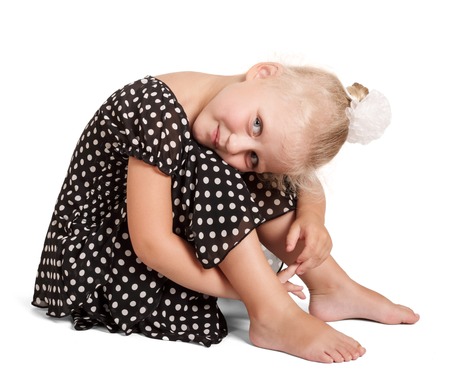 A cute little blond girl in a dark dress with polka dots with a bow on her head sitting isolated on white background.の写真素材
