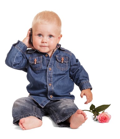 Cute little boy in a blue shirt and jeans sitting holds a mobile phone, a rose isolated on a white background.の写真素材