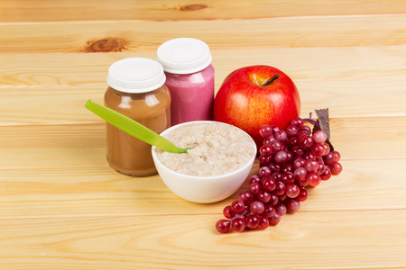 A bowl of oatmeal and jars with fruit puree, apple, bunch of grapes on a background of light wood.の写真素材