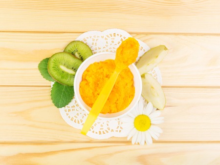 A bowl and a spoon of pumpkin puree, apple slices, kiwi on light wood background. View from above.の写真素材