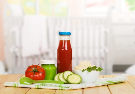 A bottle of tomato juice, vegetable puree bank, bowl with cauliflower, squash, cotton napkin and spoon on a background of the kitchen.の写真素材